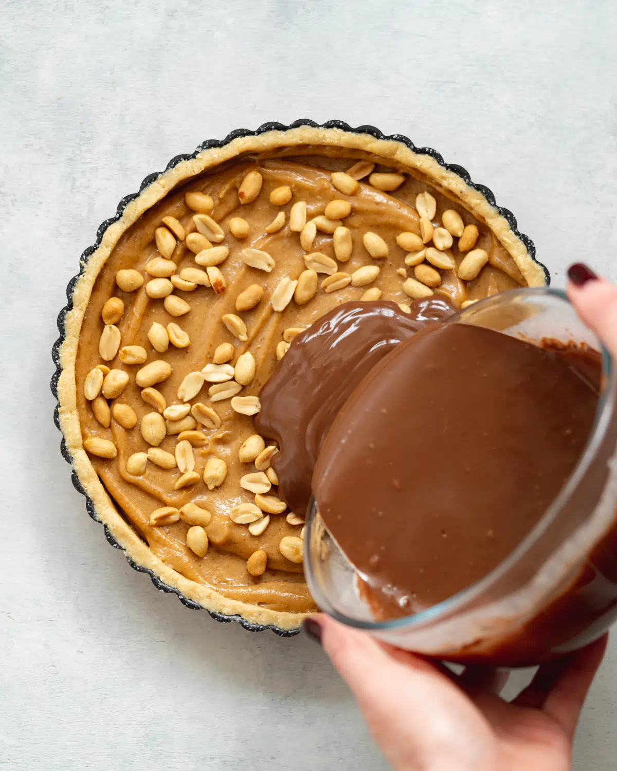 pouring melted chocolate ganache into a tart with peanut butter caramel in it.