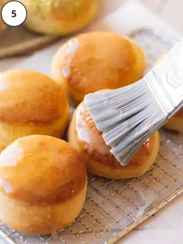a pastry brush with condensed milk being used to glaze donuts fresh from the oven on a wire rack.
