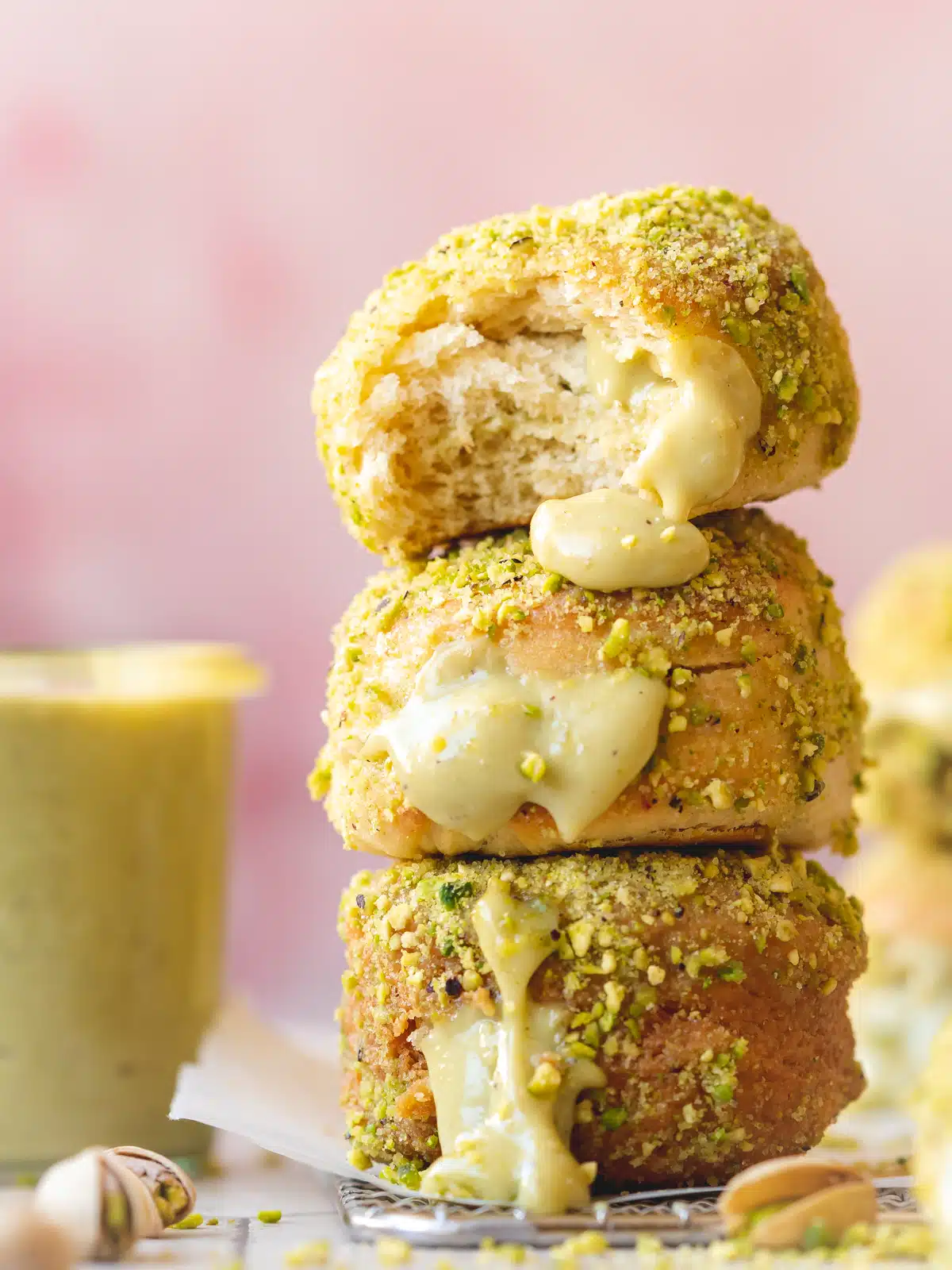 a stack of vegan yeast donuts coated in sugar and pistachios with pistachio cream oozing out from their centers. The background is pink which contrasts with the small green jar of pistachio pastry cream next to the doughnut pile.