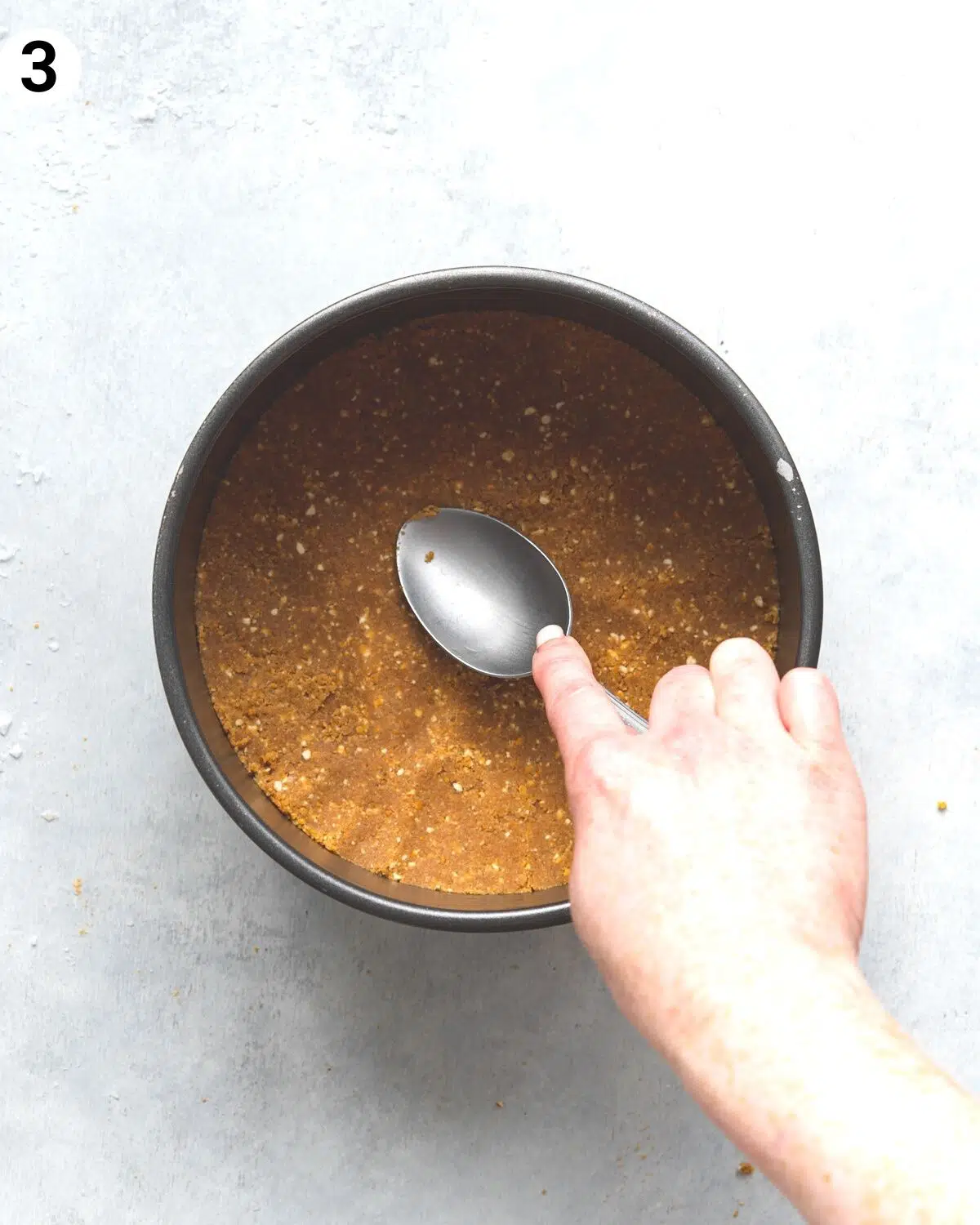 hand using a spoon to press the cookie base into a cake tin.
