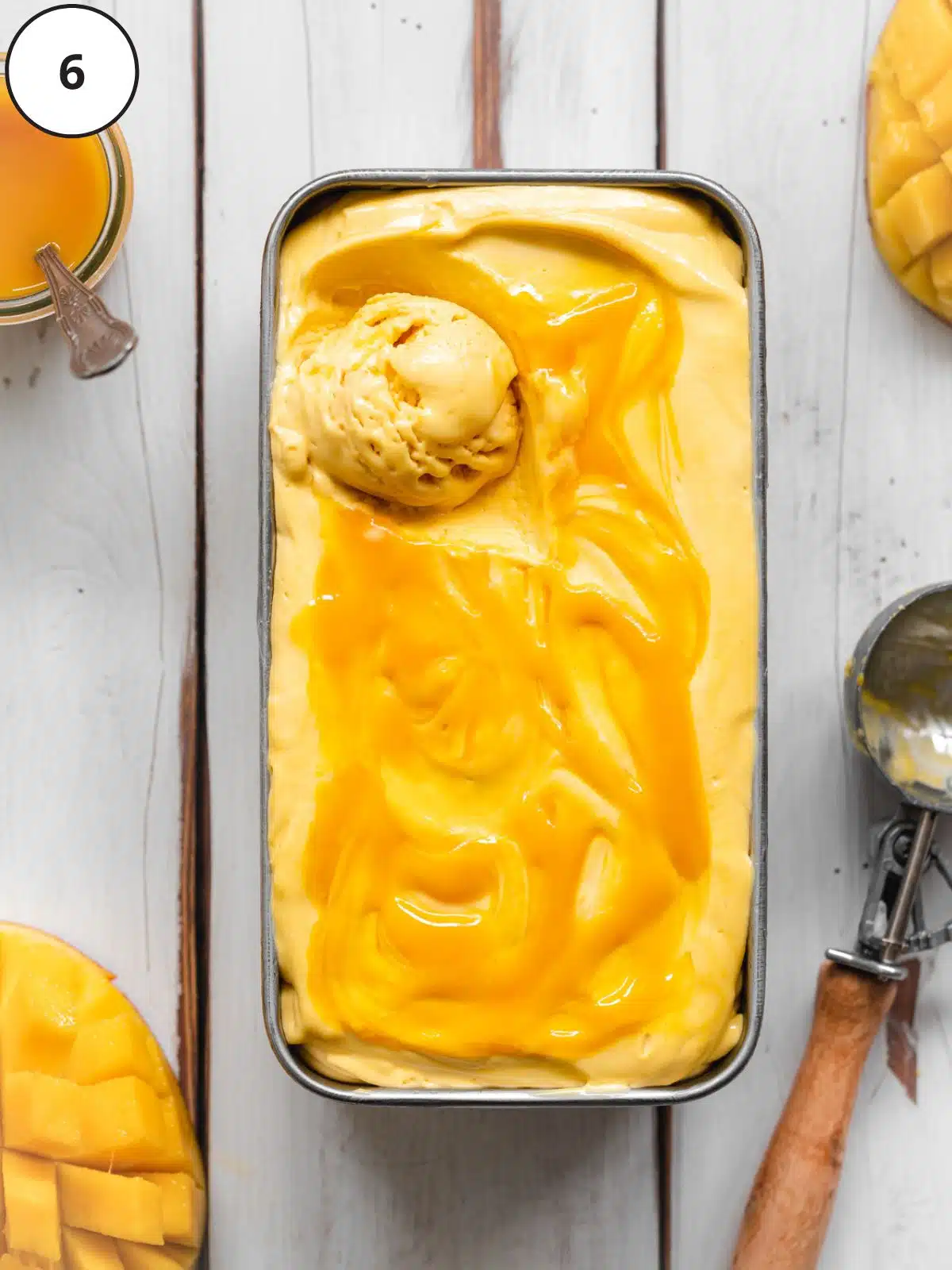 freshly homemade mango ice cream in a loaf pan with mango puree running through it, there are fresh mangoes scattered around.