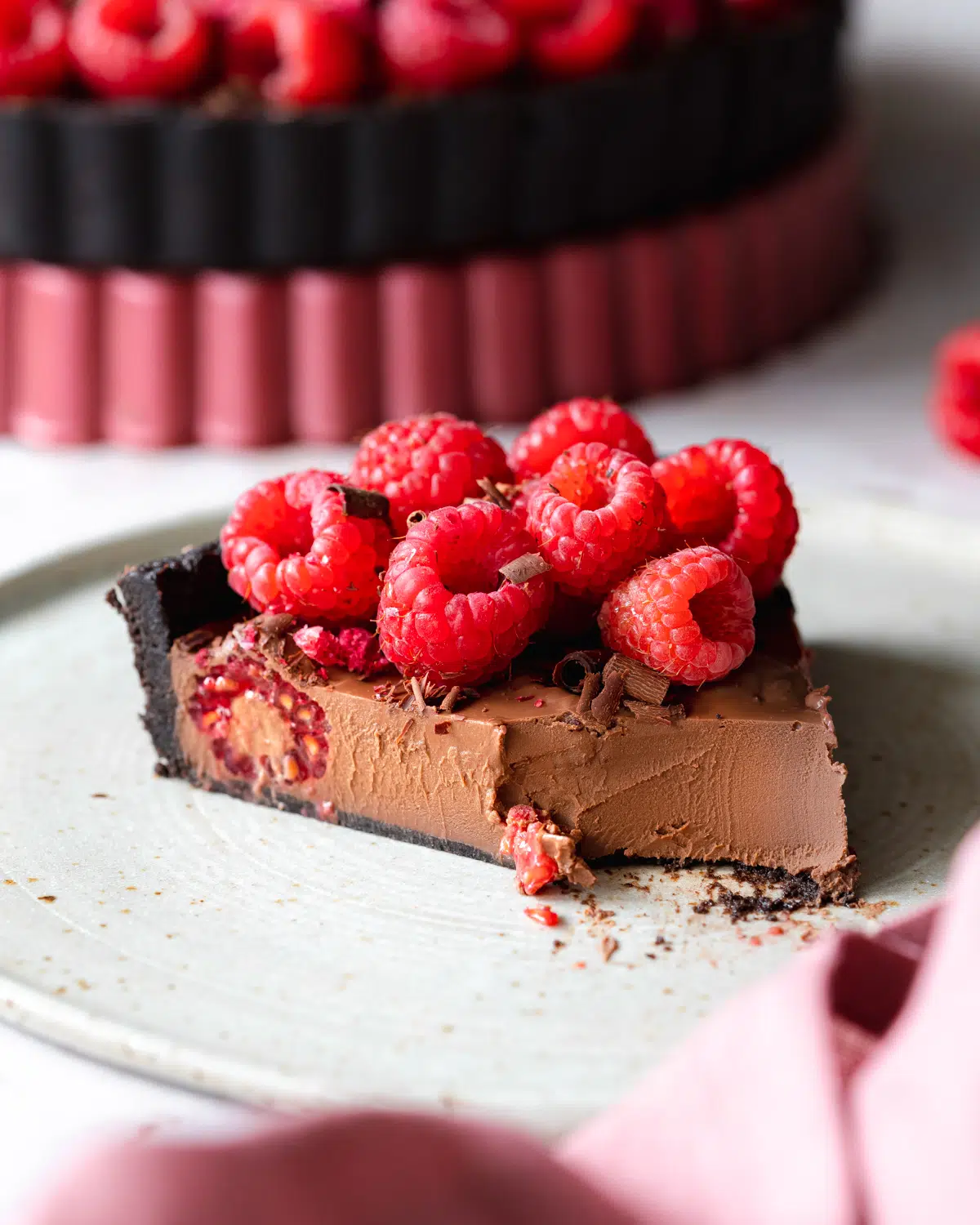 slice of chocolate ganache tart with raspberries on top, and pink linen in the foreground.