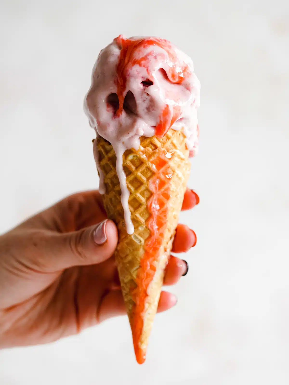 hand holding up a strawberry ice cream cone with strawberry sauce dripping from it.