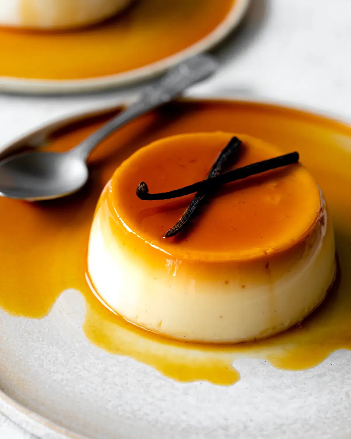 caramel flan on a plate with caramel sauce and vanilla pods on top and spoonful taken out of it.