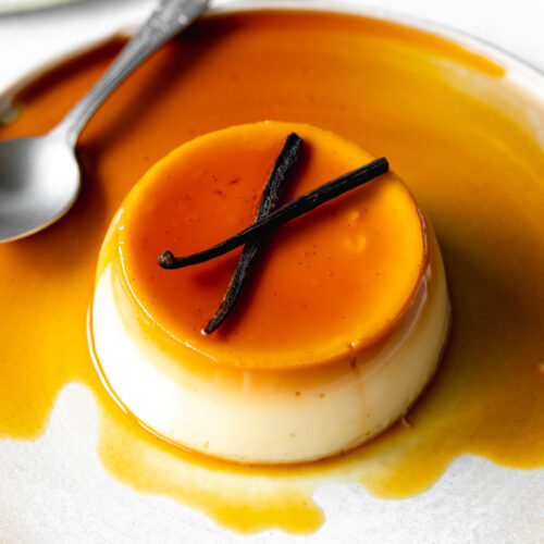 caramel flan on a plate with caramel sauce and vanilla pods on top.