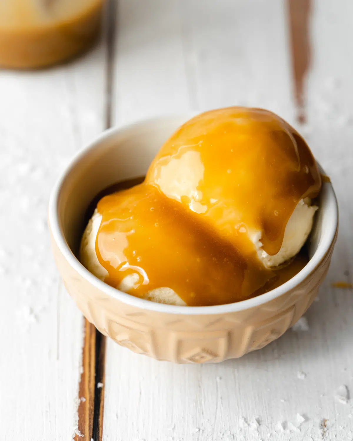 small bowl with two scoops of ice cream covered in homemade caramel sauce, on a white wooden surface.