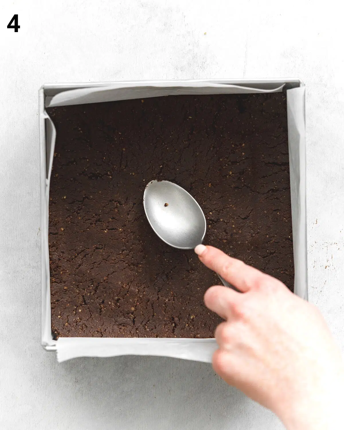top down view of hand pressing down brownie base with a spoon into a square can pan.