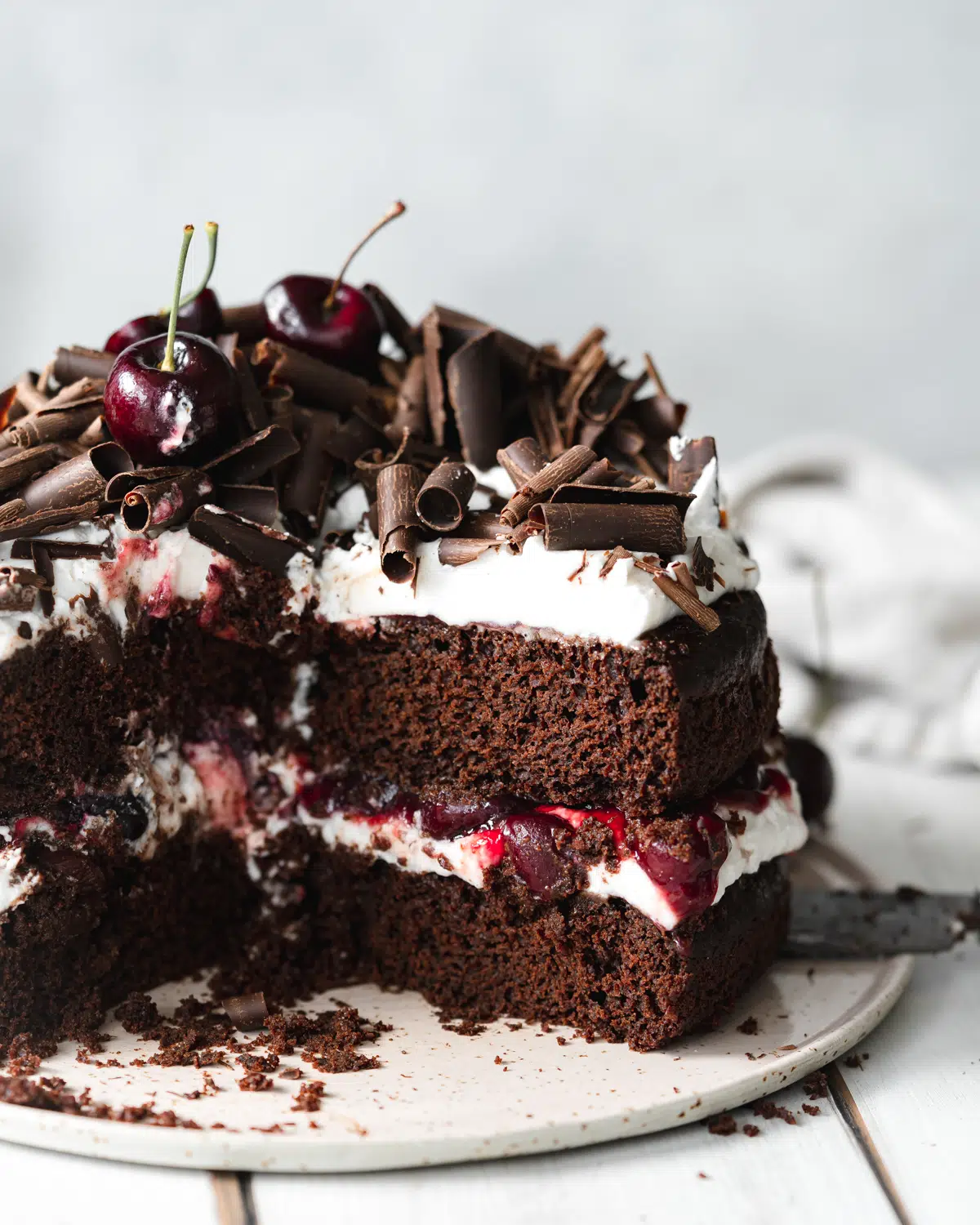 layered black forest cake with chocolate curls and cherries on top, on a white wooden surface.
