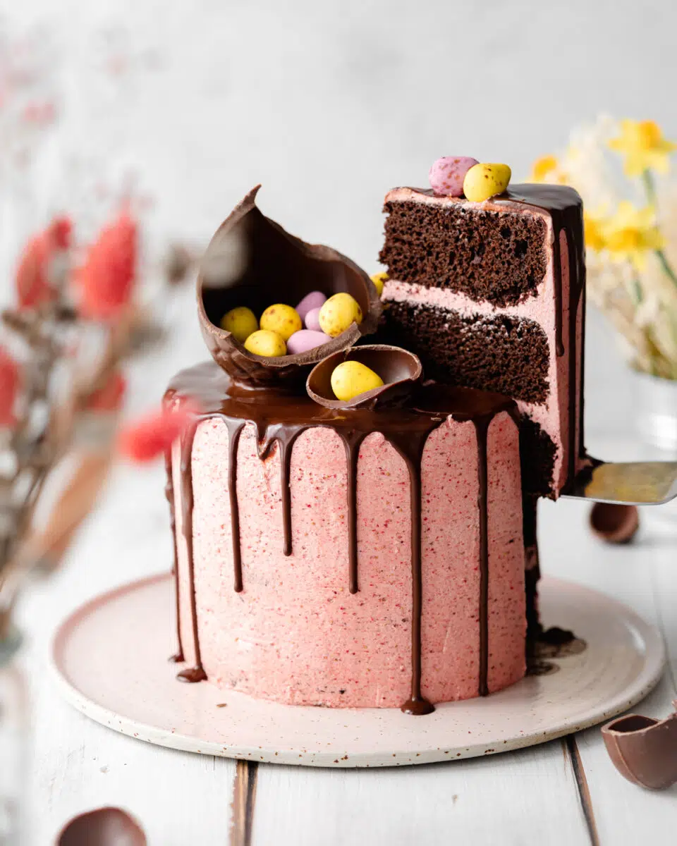pink layer cake with chocolate drip and easter eggs and mini eggs on top on a ceramic plate.