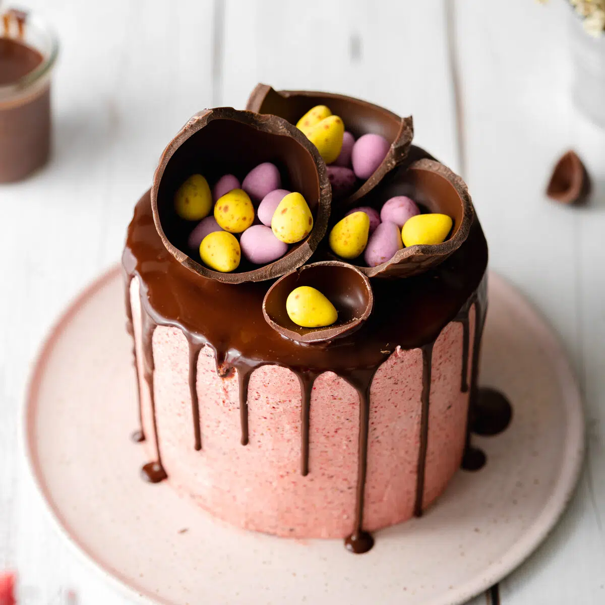 pink layer cake with chocolate drip and easter eggs on top on a ceramic plate.