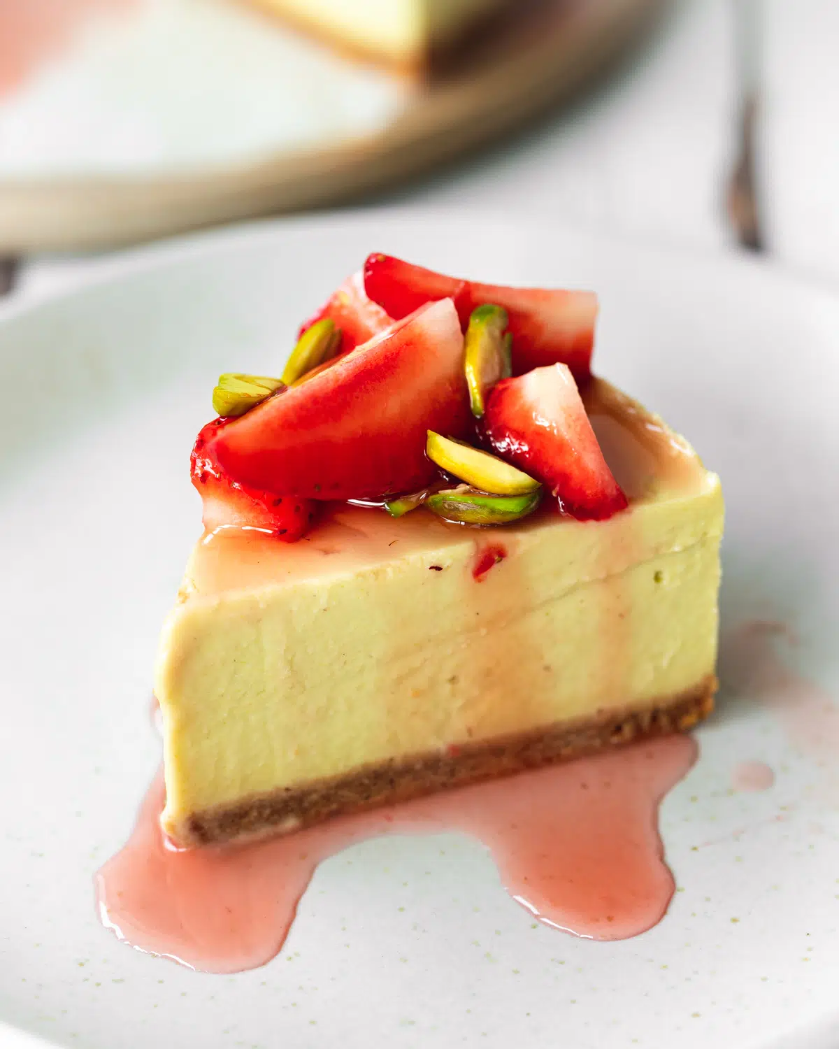slice of pistachio cheesecake with strawberries and syrup on a grey plate.
