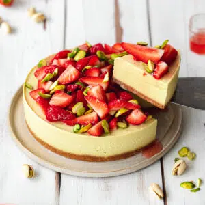 pistachio cheesecake topped with fresh strawberries and pistachio slivers on a white wooden surface.