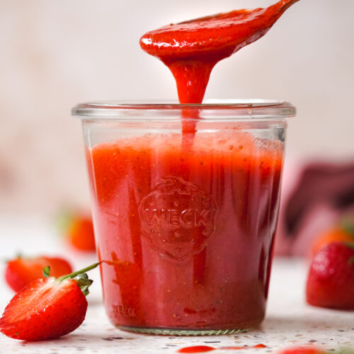 strawberry coulis in a glass jar with strawberries scattered around.