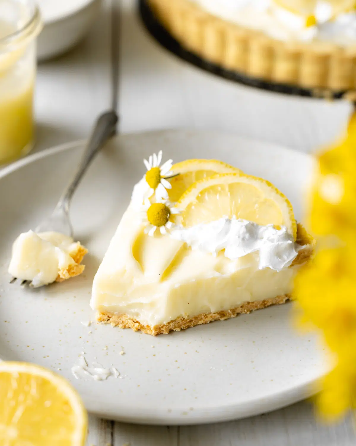 slice of creamy lemon tart with cream, lemon slices and chamomile flowers on top of a grey plate.