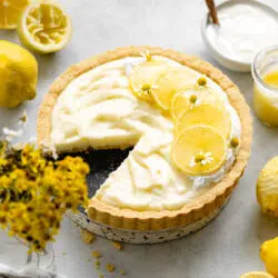 lemon tart with lemon slices on top and fresh lemons and flowers scattered around it.
