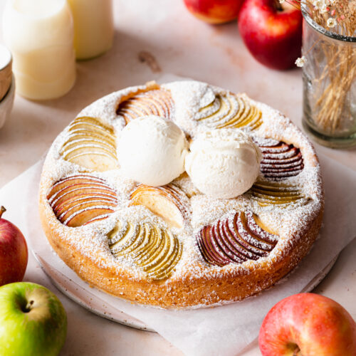 apple cake with powdered sugar and vanilla ice cream on top, and apples scattered around it.