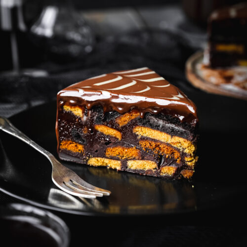 slice of chocolate biscuit cake on a black plate with a fork and chocolate ganache topping.