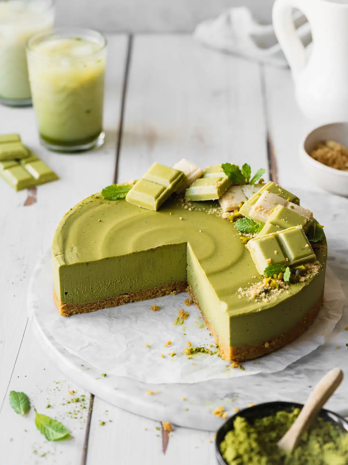 matcha cheesecake with a slice missing from it, decorated with white chocolate and matcha chocolate, with 2 matcha lattes in the background.
