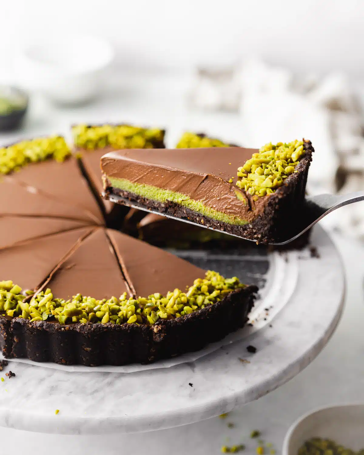 chocolate pistachio tart on a marble cake stand with a slice being lifted with a cake slice.