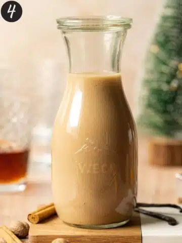 eggnog in a glass bottle with spices around it and a small christmas tree in the background.