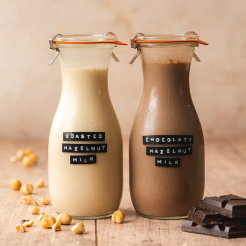 a jug of hazelnut milk next to a jug of chocolate milk with nuts and chocolate scattered around them.
