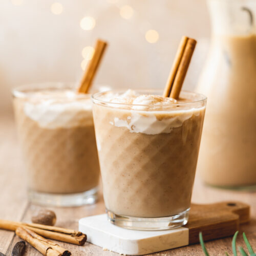 two glasses of eggnog with a cinnamon stick and whipped cream on top of each of them.
