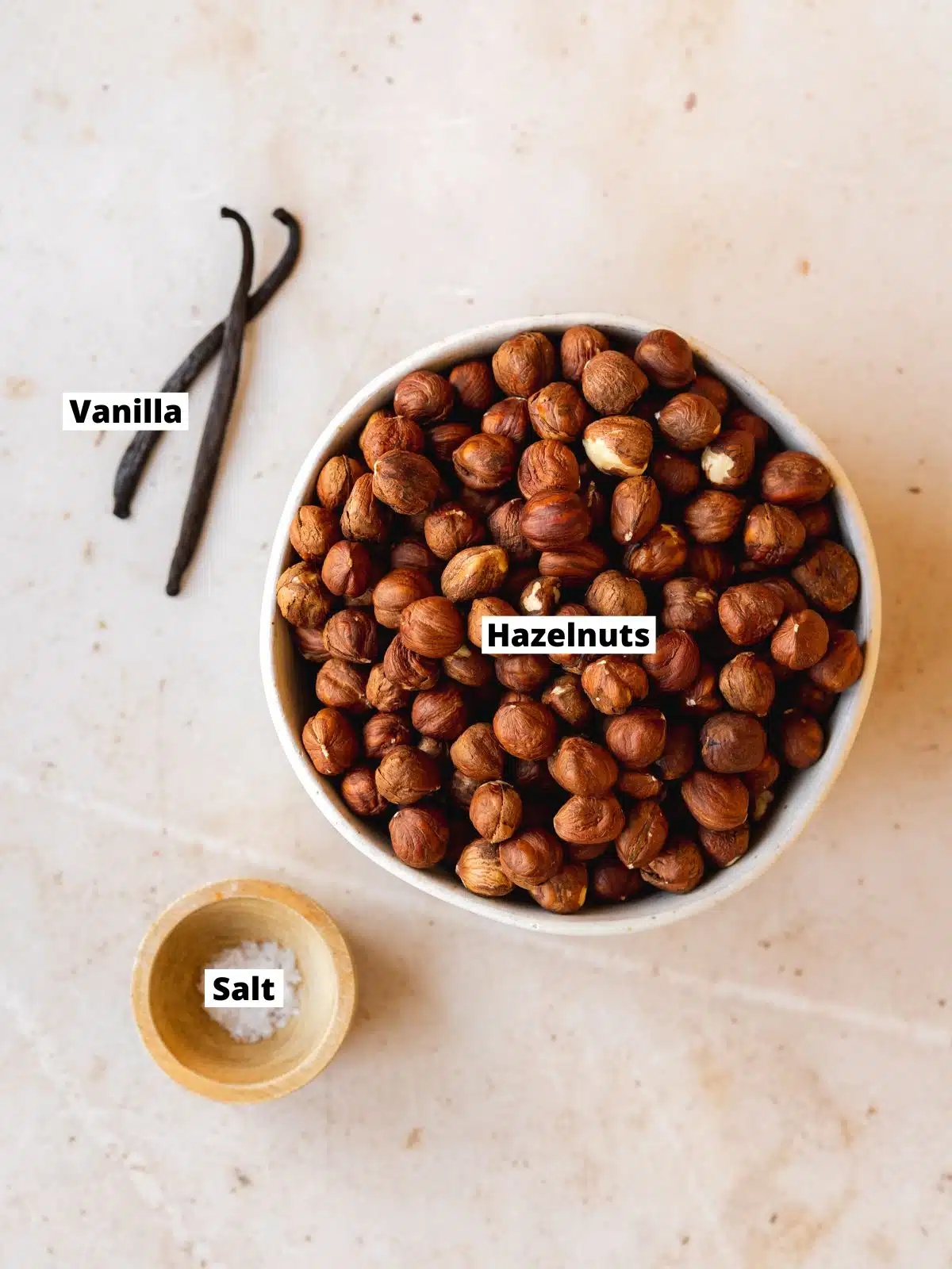 hazelnuts in a bowl next to a pinch bowl of salt and 2 vanilla pods.