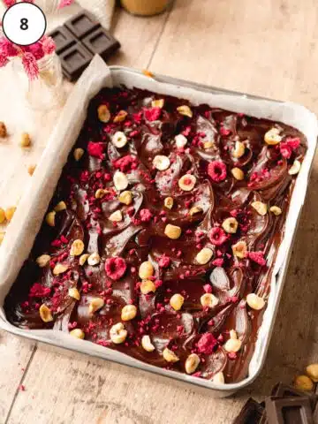 no bake brownies in a parchment lined tray with ganache, hazelnuts, and freeze-dried raspberries on top.