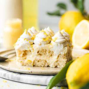 a slice of vegan limoncello tiramisu on a ceramic plate with leafy fresh lemons, lemon curd, milk and limoncello in the background.