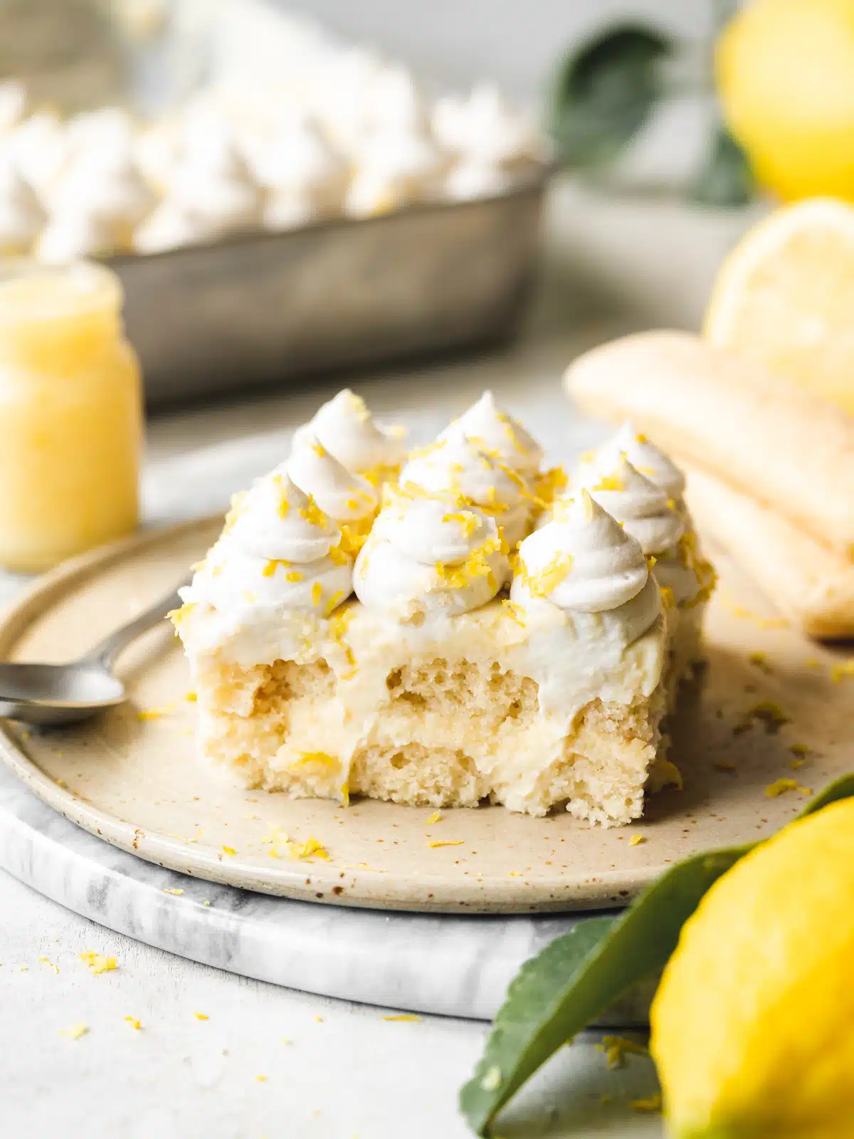 a slice of vegan lemon tiramisu with whipped cream topping on a ceramic plate with lemon curd and lemons in the background.