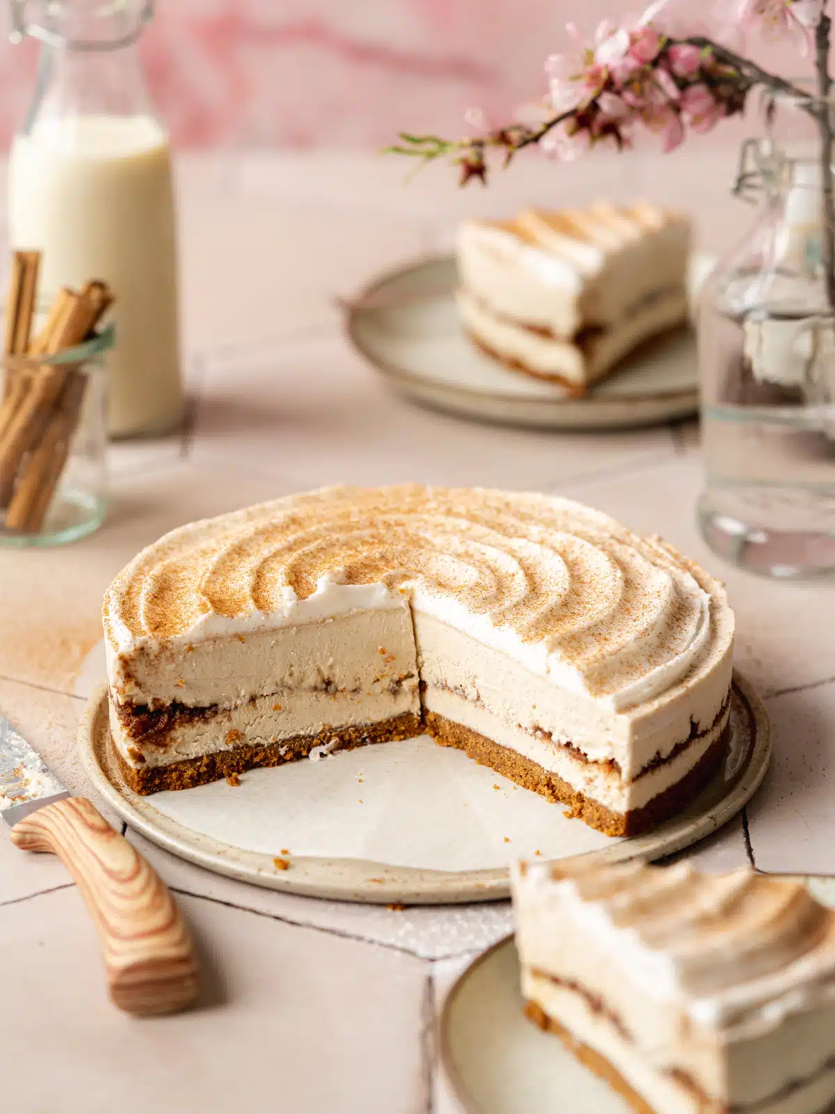 45 degree angle shot of vegan cinnamon roll cheesecake with two slices taken out, one placed on a plate in the foreground and one on a plate in the background with a glass jug of nut milk to the side.