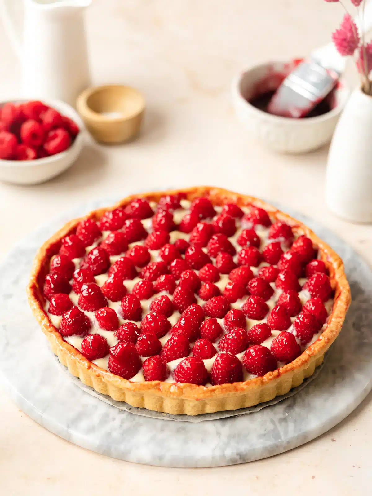 vegan french raspberry tart filled with dairy-free pastry cream and topped with raspberries on a marble surface and a bowl of fresh raspberries in the background.