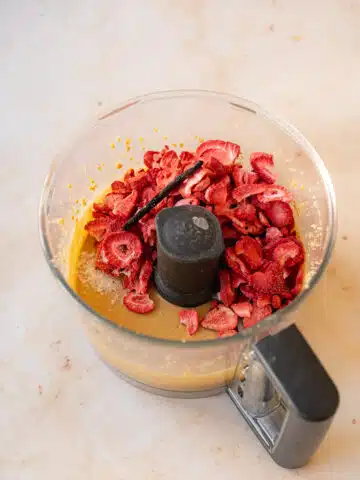 Creamy peanut butter in the pitcher of a food processor with freeze-dried strawberry slices and half a vanilla pod added.