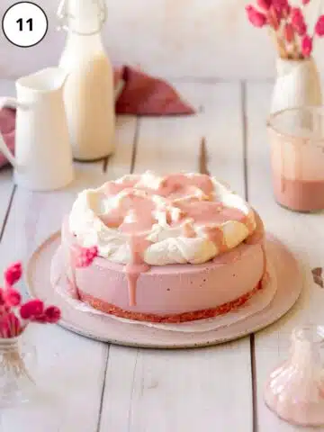 pink vegan cheesecake with whipped cream and strawberry sauce on top on a white wooden surface.