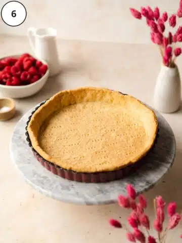 vegan pastry shell in a tart tin sitting on a marble cake stand with fresh raspberries and pink flowers in the background.