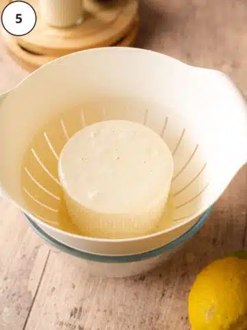 round of ricotta cheese in a colander on top of a straining bowl.