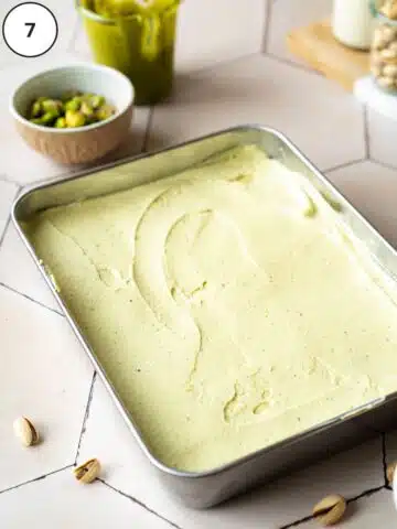 a smooth layer of dairy-free pistachio mascarpone in a rectangular baking pan. There is a small jar of pistachio butter and a bowl of pistachios in the background.