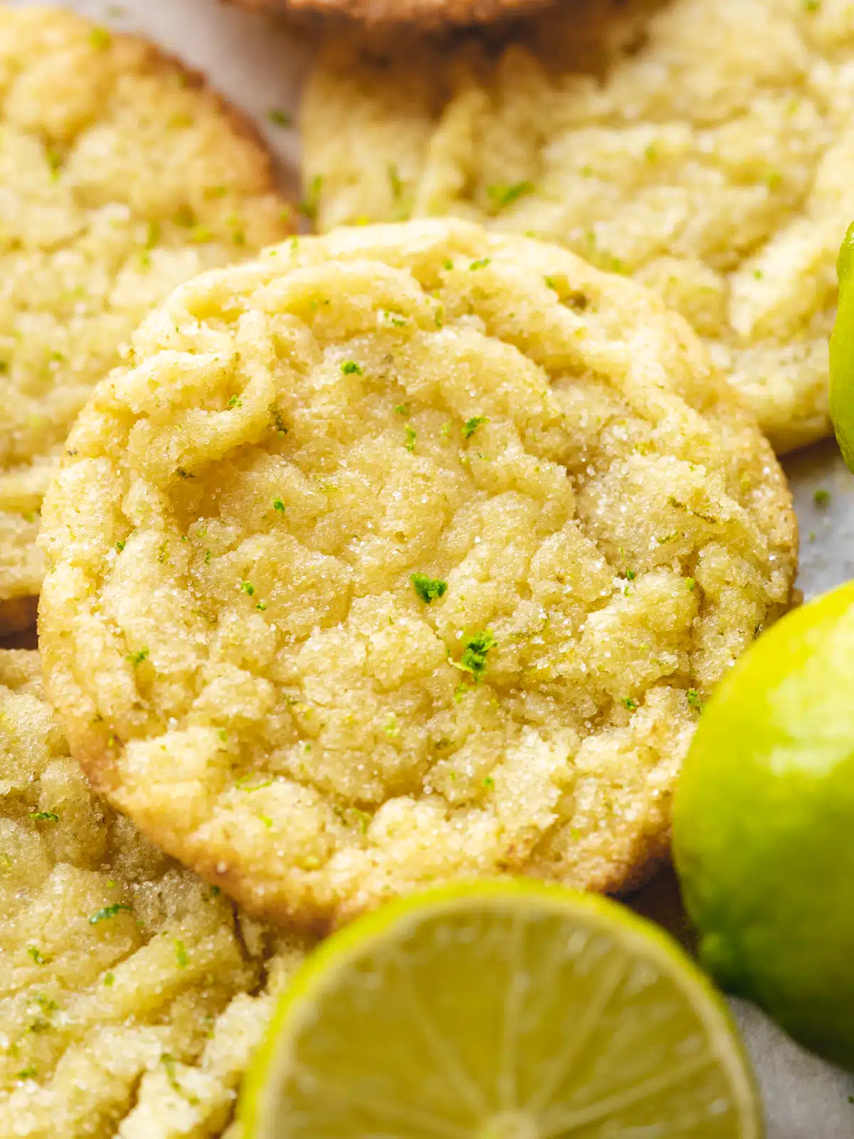 Closeup shot of a sugar and lime-zest coated vegan key lime cookie with a halved lime in the foreground and a pile of more cookies blurred out in the background.