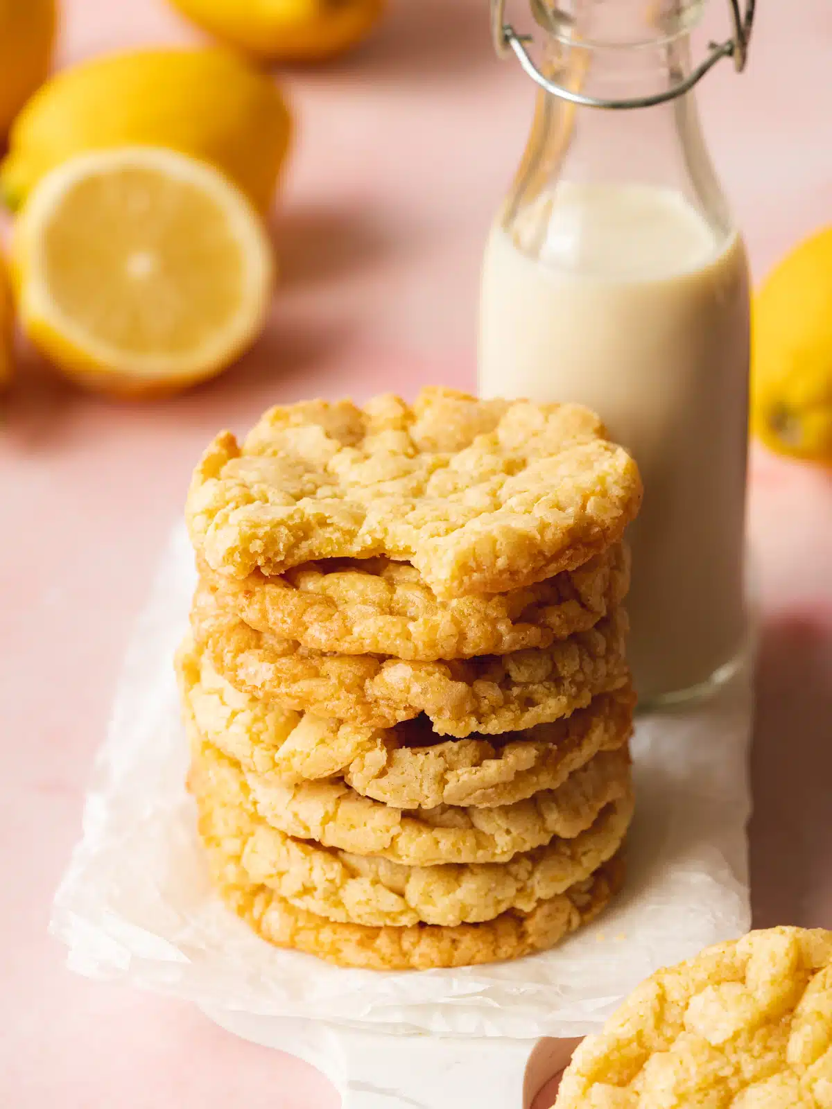 a stack of vegan lemon cookies on a sheet of baking paper with a bottle of milk next to them. There are fresh lemons in the background and the yellow is contrasting against the pink surface.