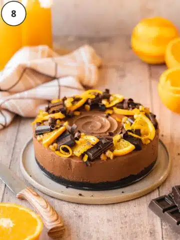 Chilled orange chocolate cheesecake removed from the springform tin and decorated with wedges of candied orange, strips of candied orange zest, and vegan dark chocolate shavings.