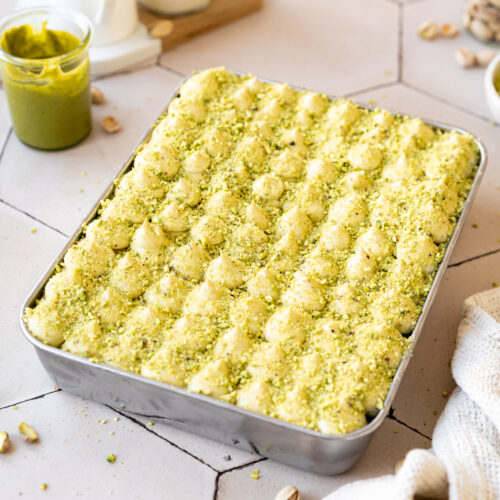 pistachio tiramisu in a rectangular cake tray with ground pistachios on top and a small jar pf pistachio butter in the background.