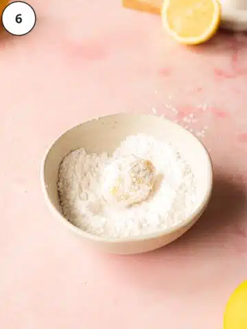 a lemon cookie being rolled in a bowl of powdered sugar.