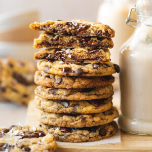 tall stack of the best vegan chocolate chip cookies sprinkled with flaky sea salt sitting next to a glass jug of homemade nut milk.