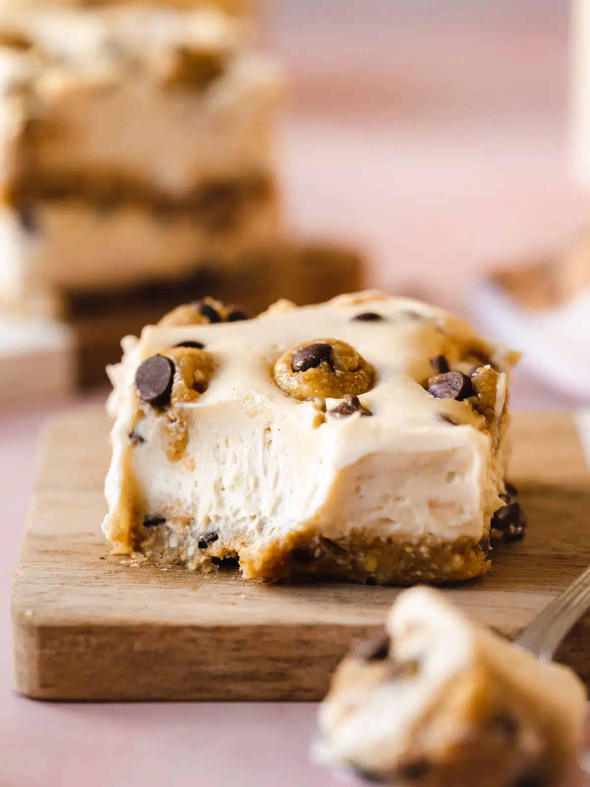 One vegan chocolate chip cookie dough cheesecake bar on a wooden plate with a bite taken out to show the creamy consistency.