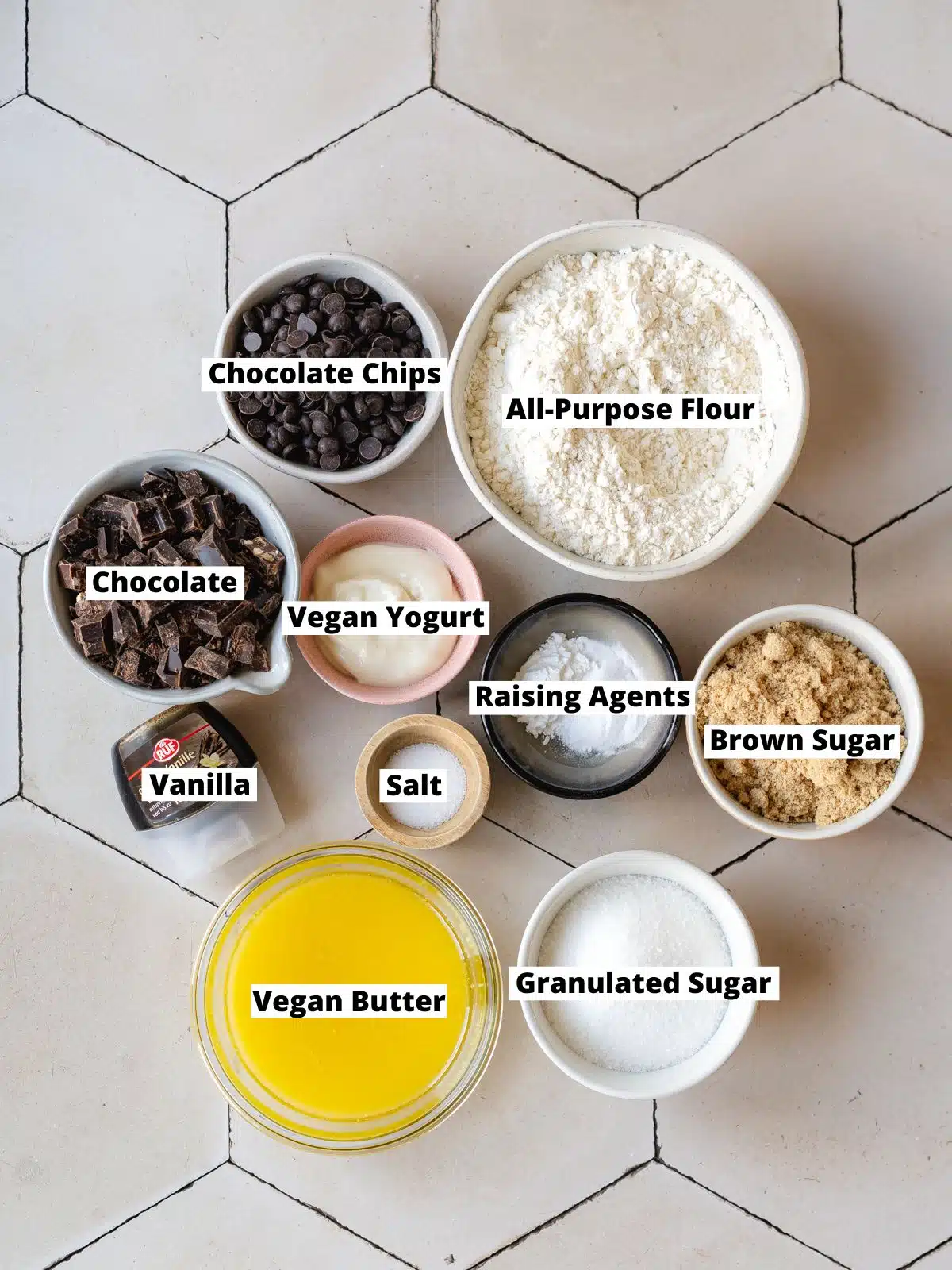 ingredients needed to make the best vegan chocolate chip cookies measured out into bowls on a white table with hexagonal tiles.