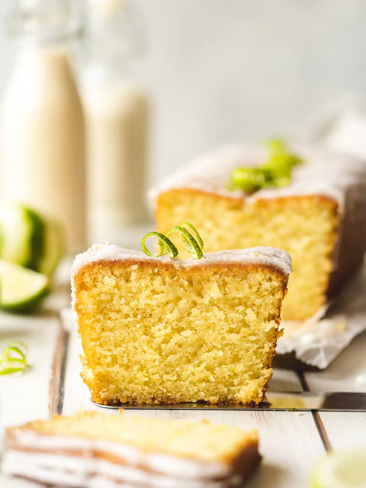 Side on hero shot of a slice of key lime drizzle cake on a cake server with the rest of the loaf cake, a few jugs of vegan milk, and quartered limes slightly blurred out in the background.