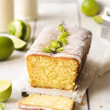 square hero image of a loaf of key lime pound cake garnished with a lime drizzle and curls of fresh lime zest. A slice is removed to show the tnder interior crumb and lightly golden crust.