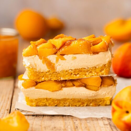 Two peach cheesecake bars stacked atop one another on top of a piece of parchment with some whole peaches scattered about the scene.