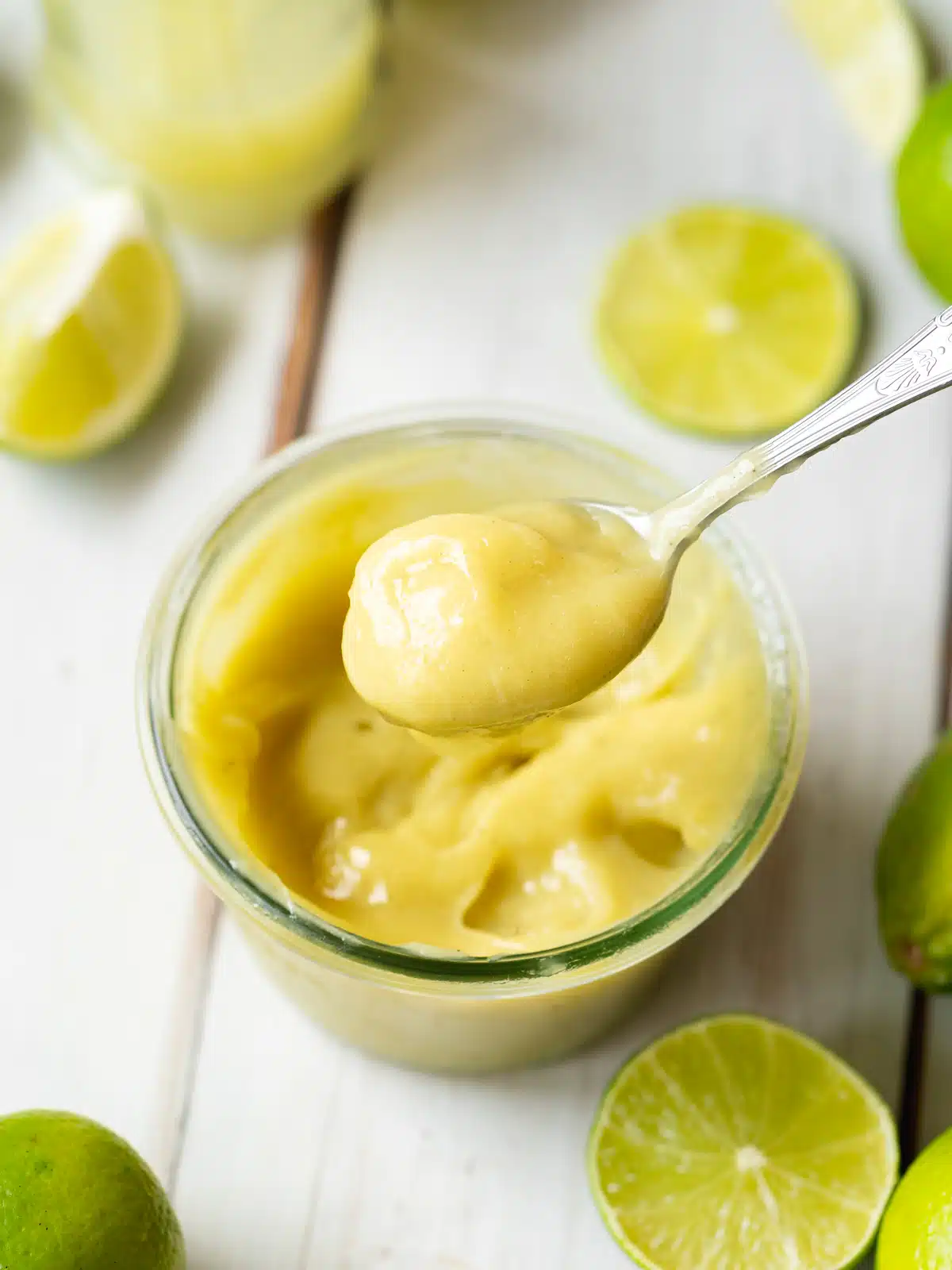 thick lime curd without eggs being spooned out of a jar. It has a vibrant green color and there are fresh lime slices scattered around it.