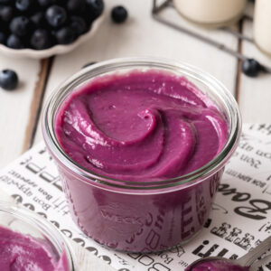 smooth and creamy blueberry curd in a glass jar on top of newspaper with fresh blueberries in the background.